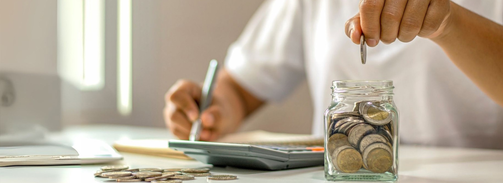 Une main laissant tomber une pièce dans un bocal plein de pièces. D'autres pièces sur une table et une calculatrice, calculs pour économiser sur la flotte. Réduire les dépenses de carburant